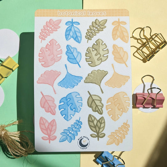 Sticker sheet with various illustrated leaves in bright colours, on a green and yellow background with stationery strewn about.